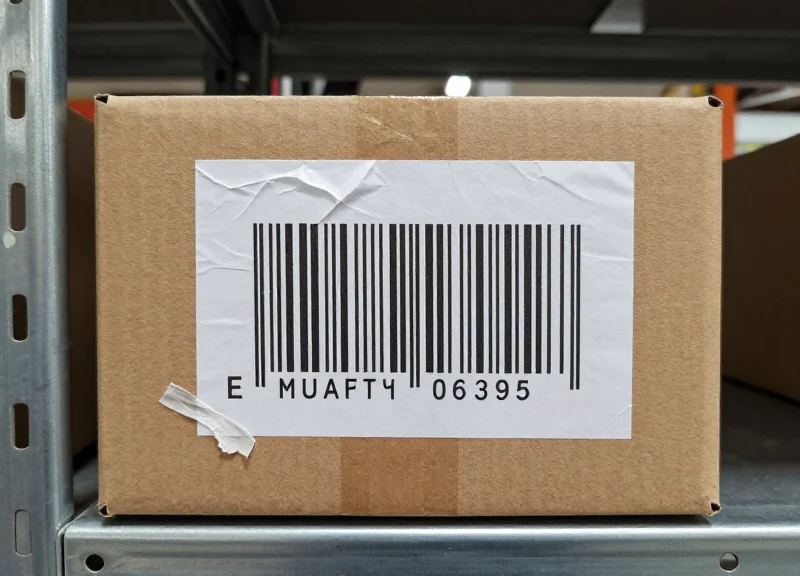 Close-up of a damaged cardboard package with a peeling barcode label in a warehouse setting.